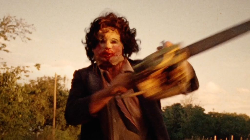 A masked man wildly swings a chainsaw outdoors in a chaotic and terrifying scene from The Texas Chain Saw Massacre.