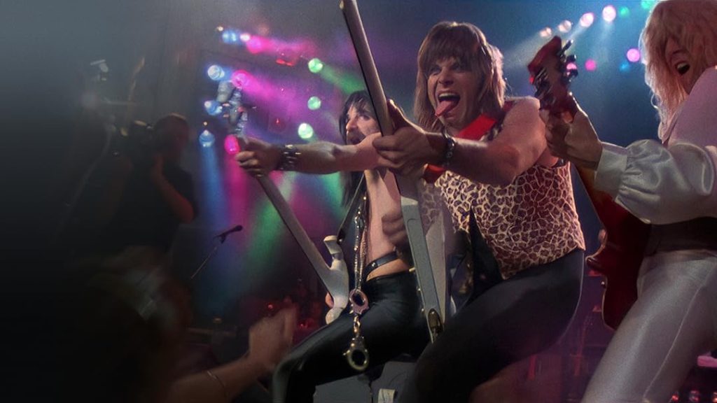 A rock band performs energetically on stage under colorful concert lights while musicians lean toward the crowd playing guitars in This Is Spinal Tap.