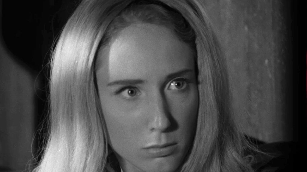 A terrified blonde woman stares ahead with wide fearful eyes in a black-and-white scene from Night of the Living Dead.