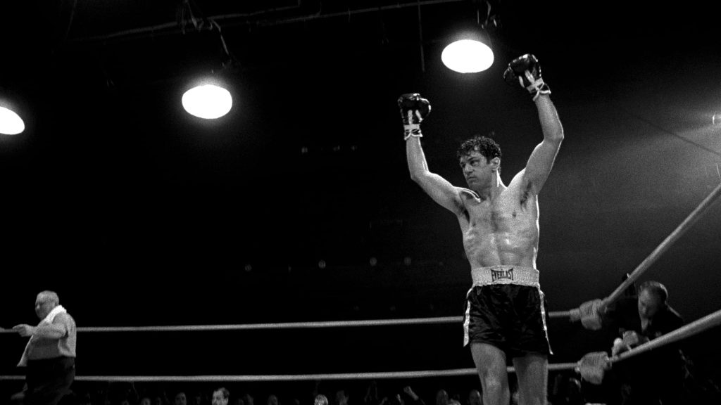 A boxer stands triumphantly in a black-and-white boxing ring with both gloved fists raised under bright overhead lights in a scene from Raging Bull.