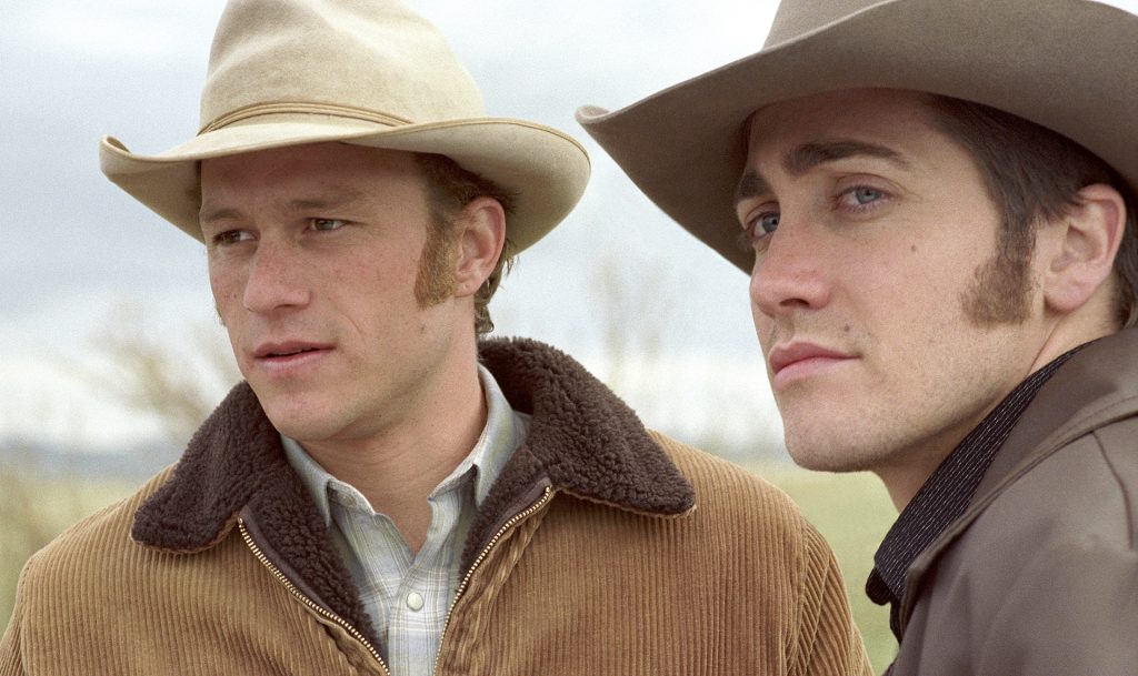 Two cowboys in wide-brimmed hats stand outdoors in a quiet rural landscape in a scene from Brokeback Mountain, both looking off into the distance with serious expressions.
