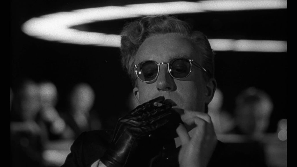 A black-and-white close-up of a man wearing round sunglasses and a black glove sits under circular overhead lights in a tense scene from Dr. Strangelove.