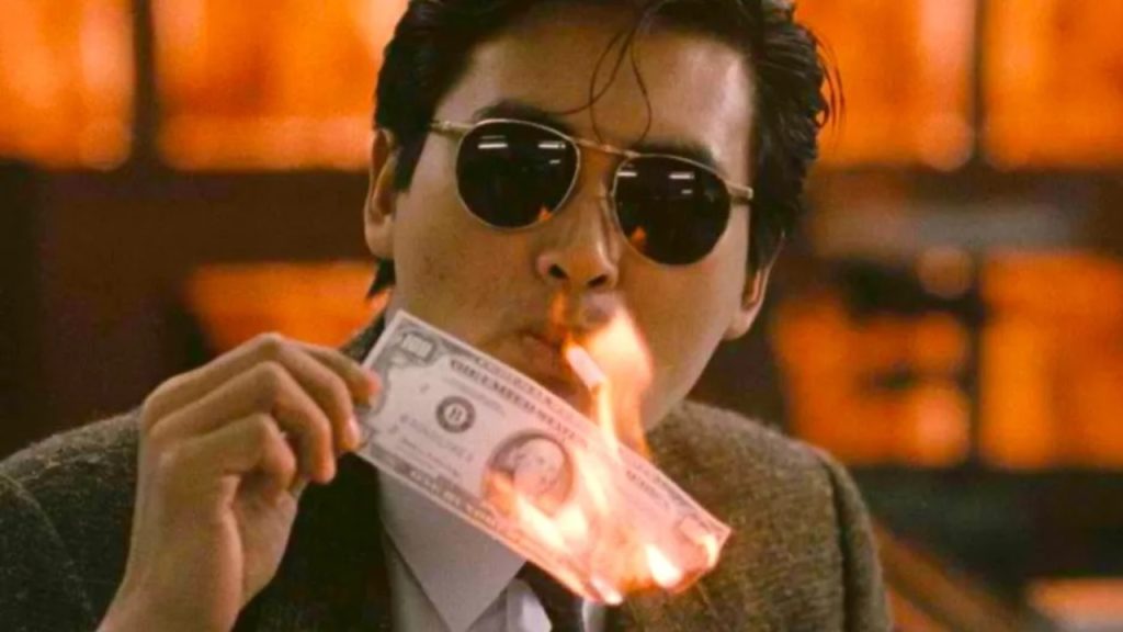 A man wearing dark sunglasses calmly lights a cigarette with a burning banknote in a stylish action scene from A Better Tomorrow.