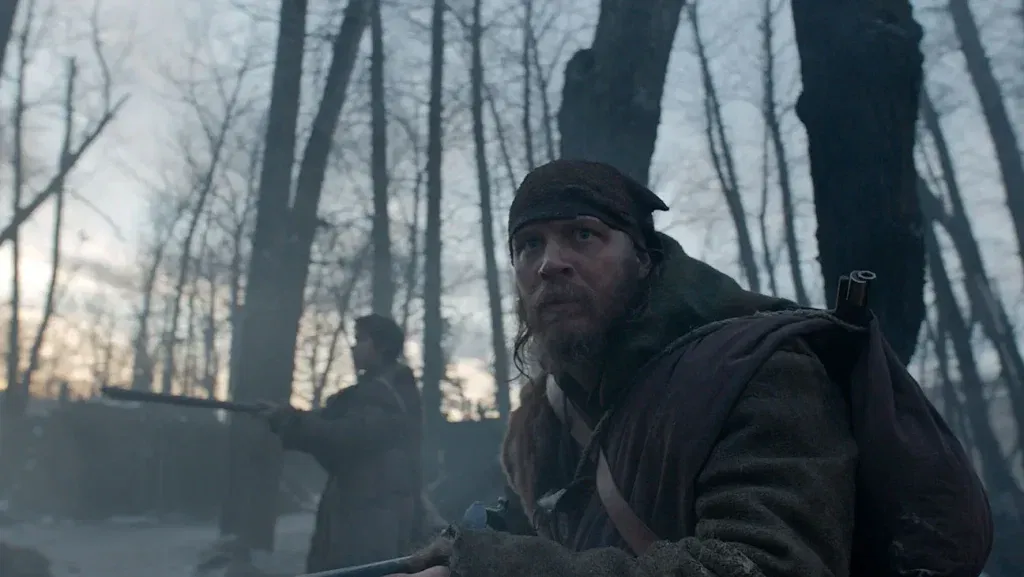 Tom Hardy as John Fitzgerald, bearded and wearing frontier clothing and a dark headband, crouching in a misty winter forest with a silhouetted figure and bare trees behind him in The Revenant (2015)