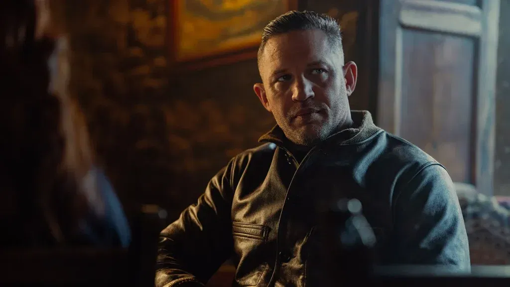 Tom Hardy as Harry Da Souza, wearing a leather jacket over a grey turtleneck, sitting across from someone in a dimly lit pub interior in MobLand (2025)