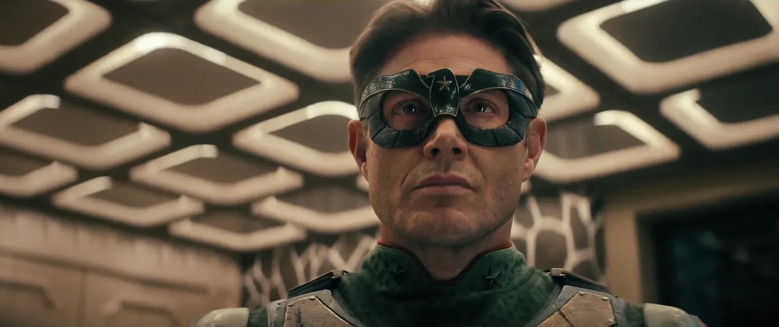 Jensen Ackles as Soldier Boy wearing a dark half-face mask with a star emblem and green tactical suit, looking upward in a retro-futuristic interior in Vought Rising (2027)