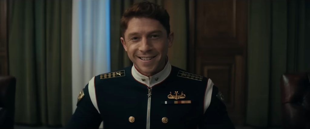 A young man with auburn hair smiling broadly in a 1950s-style military dress uniform with gold buttons and medal ribbons in Vought Rising (2027)