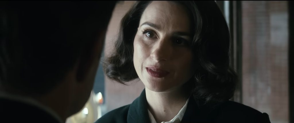 Aya Cash as Clara Vought, dark-haired in 1950s period clothing, leaning in toward another person 