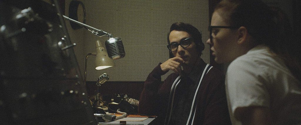 Two young people huddle together in a dimly lit 1950s radio studio, a vintage microphone and rotary phone on the desk before them, their expressions taut with concentration and unease.