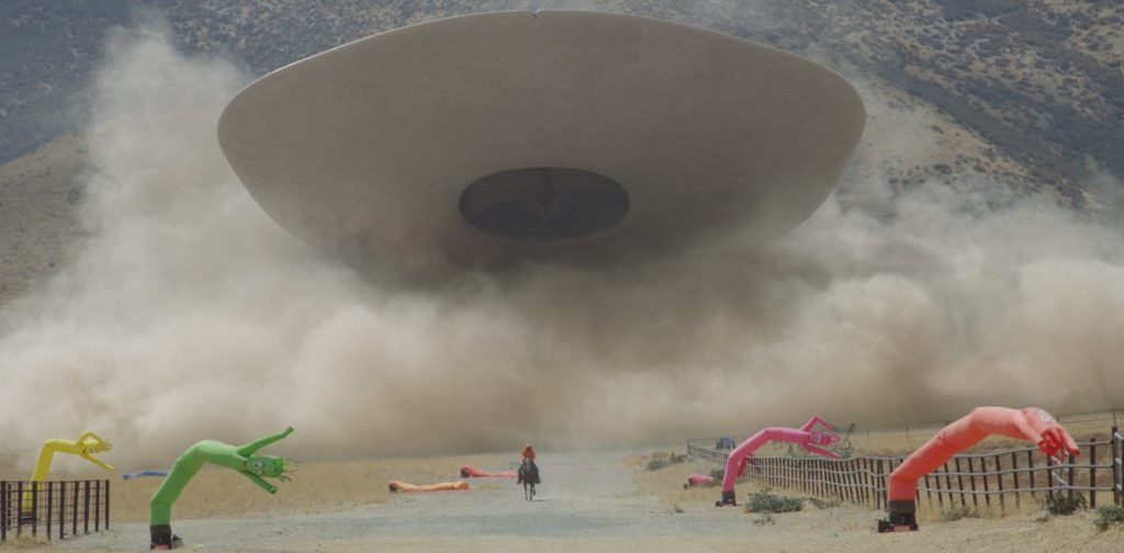 A lone rider on horseback faces a massive disc-shaped entity descending through billowing dust clouds above a California valley, flanked incongruously by colorful inflatable tube dancers whipping in the wind.