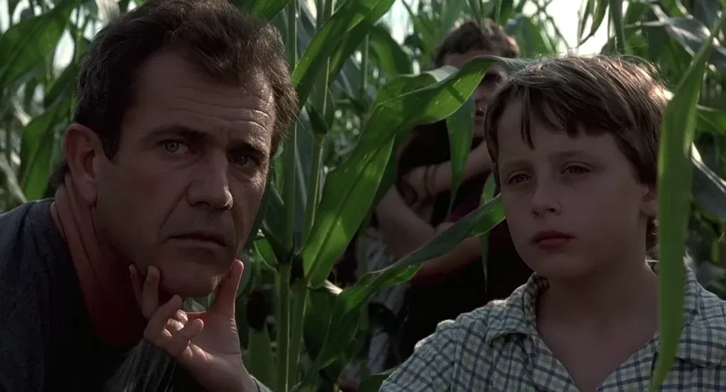 A man and a young boy crouch tensely among tall cornstalks, their faces close together, eyes wide and fixed on something unseen, with two more figures partially visible behind them.