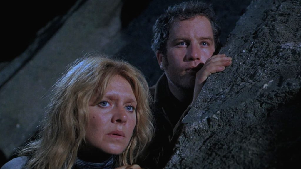 A disheveled woman and a wide-eyed man peer around the edge of a dark concrete structure, their faces lit from below by an unearthly glow, both caught between terror and pure astonishment.