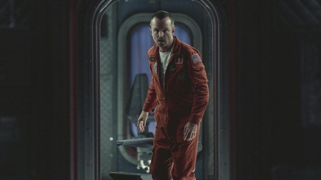 Aaron Paul stands in a futuristic spacecraft corridor wearing an orange jumpsuit in Black Mirror's Beyond the Sea episode. He will next be seen in Anything but ghosts