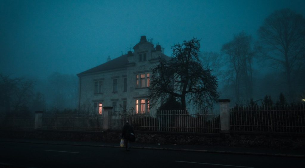 A large stone house shrouded in blue-tinted fog at dusk, its windows glowing amber from within, a lone figure walking past the iron fence on the street below, bare trees looming overhead.