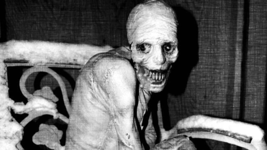 A black and white image of an emaciated, gaunt figure with sunken eyes, exposed teeth, and taut skin stretched over bone, crouched and staring directly into the camera.