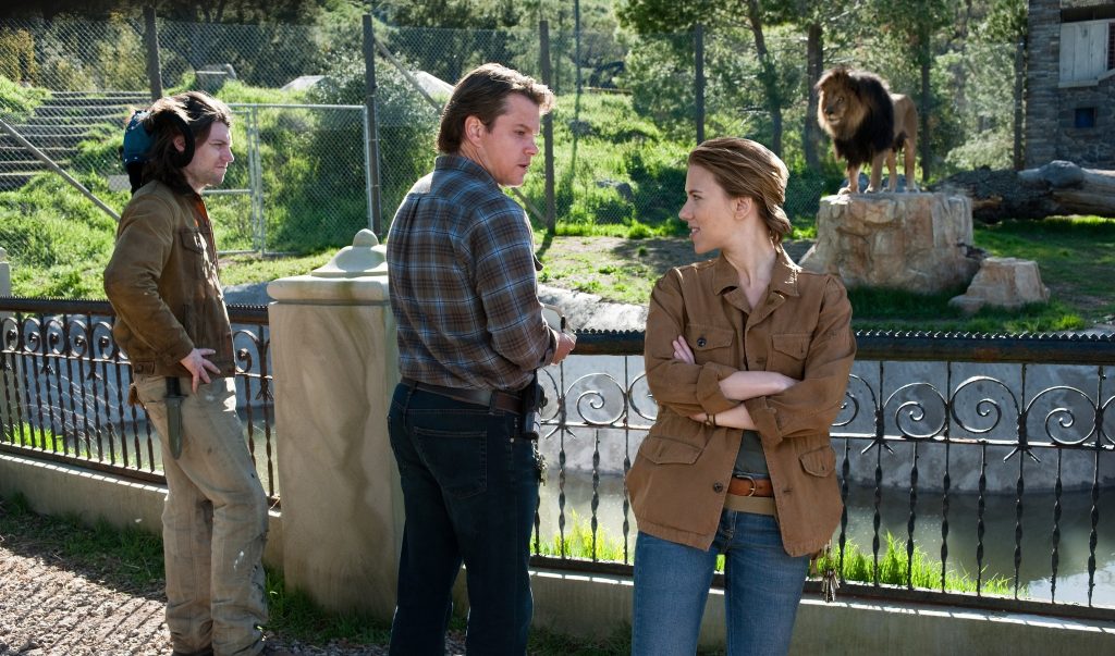 Matt Damon as Benjamin Mee and Scarlett Johansson as Kelly Foster talk at the zoo enclosure fence, with a lion standing on a rock in the background, in We Bought a Zoo