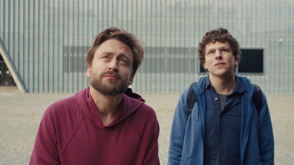 Kieran Culkin as Benji in a red hoodie and Jesse Eisenberg as David in a blue jacket stand outside a modern building in Poland, both looking upward, in A Real Pain 