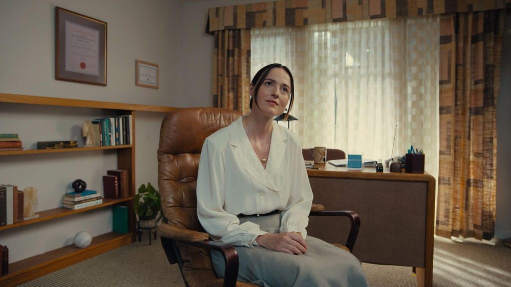 Renate Reinsve as Dr. Mary Kline seated in a leather chair in her 1990s therapist's office, surrounded by bookshelves and framed certificates, in Backrooms 