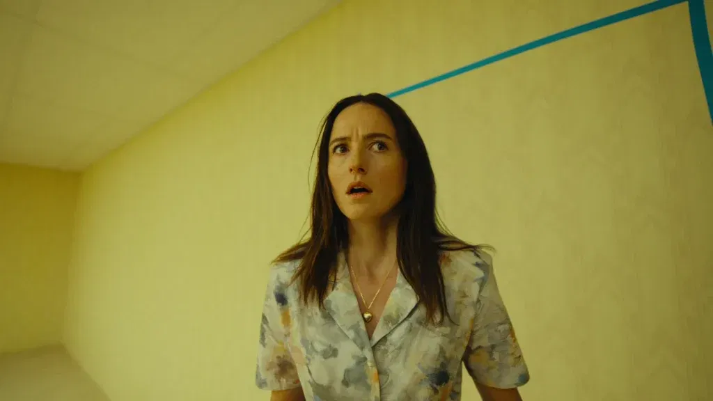 Renate Reinsve as Dr. Mary Kline looks upward in wide-eyed fear inside the yellow-walled Backrooms, a blue line marking the ceiling corner behind her in Backrooms