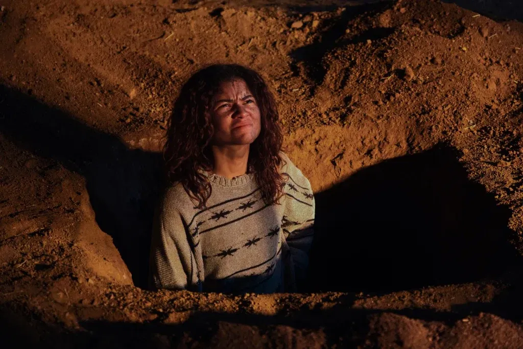 Rue stands inside a freshly dug grave at night, looking upward with a pained expression as warm light illuminates the surrounding earth.
