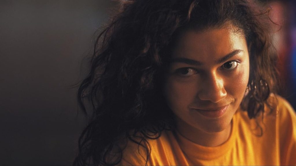 Rue looks directly toward the camera with a subtle smile, her face softly illuminated by warm ambient lighting.