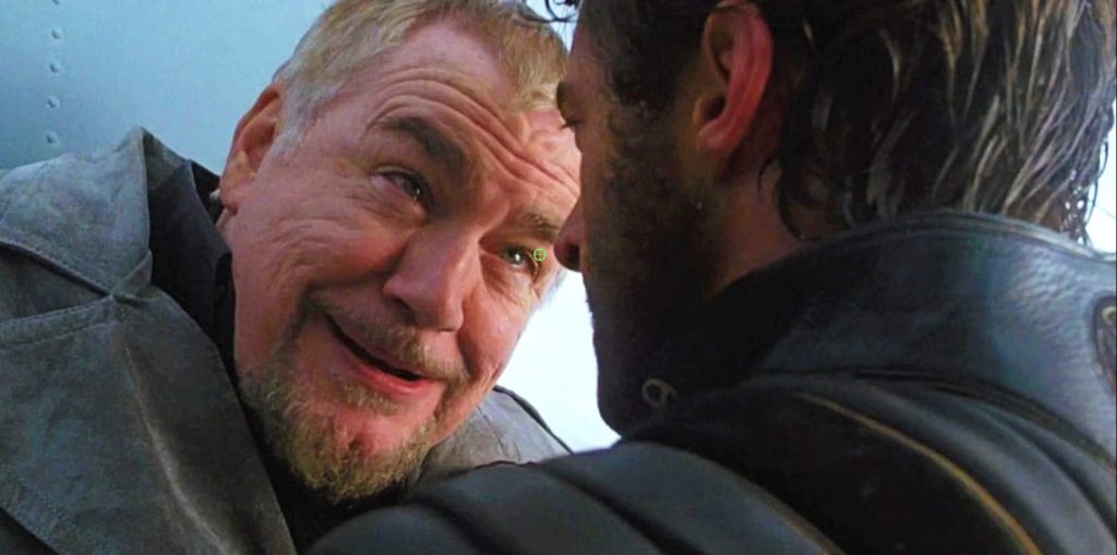 Brian Cox smiling unsettlingly close to another man's face in a tense, dimly lit scene