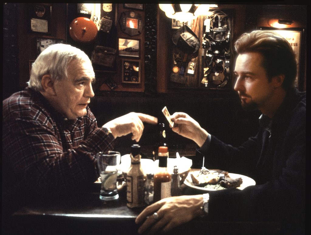 An older man and a younger man sit across from each other in a dimly lit New York bar cluttered with firefighter memorabilia, sharing a quiet, loaded moment over food and drink