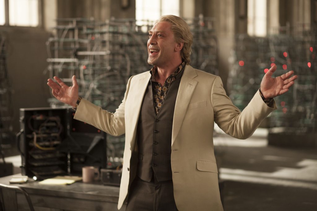 Javier Bardem as Raoul Silva in Skyfall (2012), wearing a cream-colored suit and patterned shirt while standing with his arms outstretched inside a high-tech facility, smiling confidently.