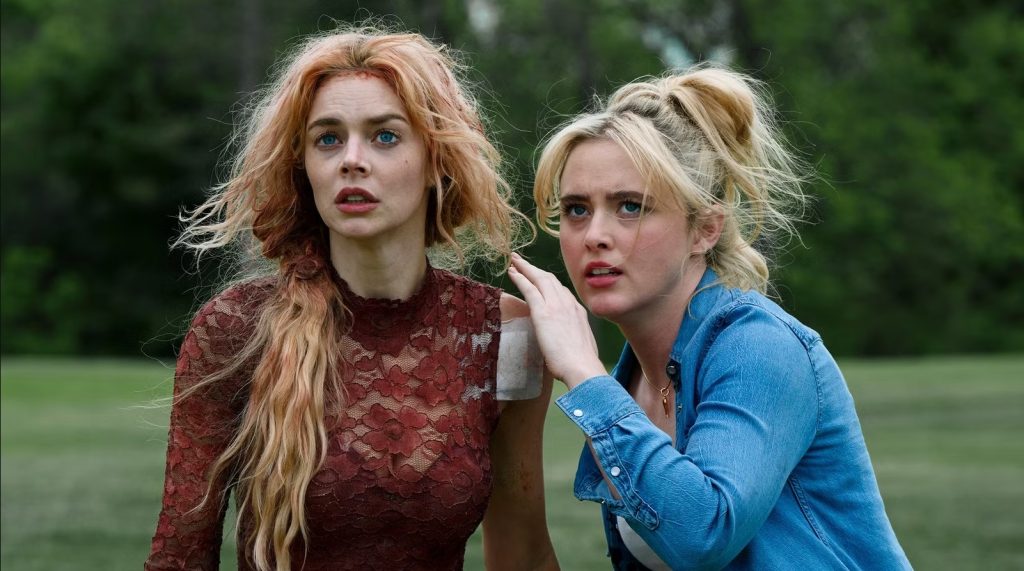Two alarmed young women outdoors, one with long auburn hair in a red lace dress with a bandaged shoulder, the other blonde in a denim shirt with her hand on the first woman's shoulder.