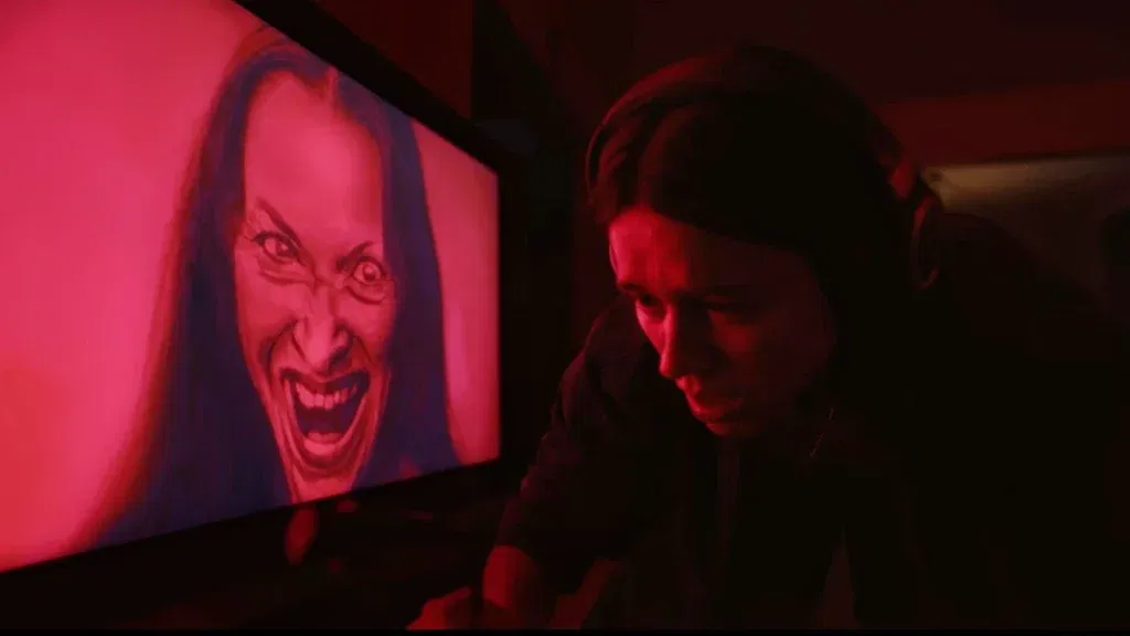 A distressed woman wearing headphones sits beside a television displaying a screaming, distorted face, bathed in intense red light.