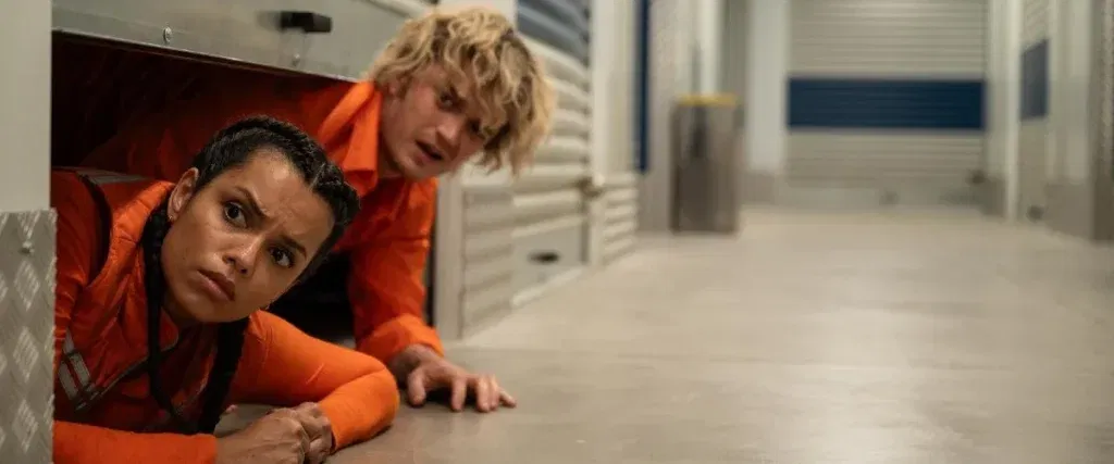 Two workers in orange uniforms cautiously crawl out of a storage unit and peer down a brightly lit corridor, looking alarmed by something off-screen.
