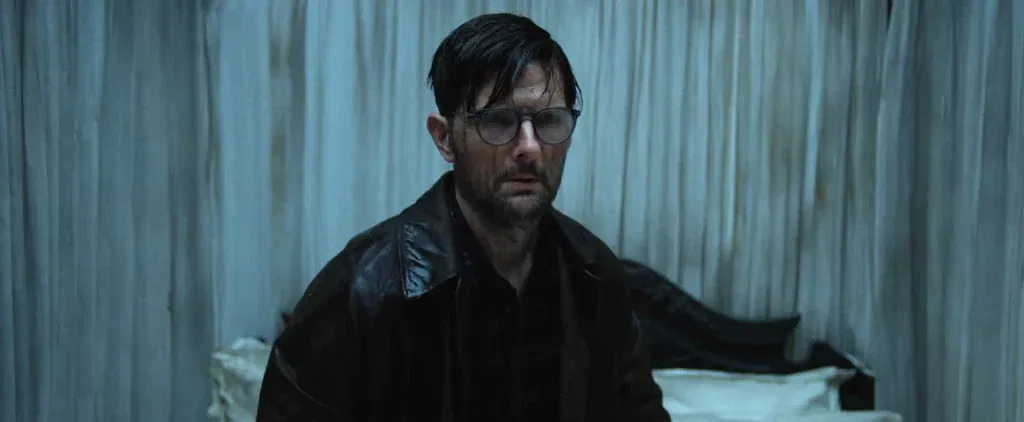 A drenched, exhausted man wearing glasses and a dark raincoat stands in a dimly lit room, staring ahead with a troubled expression.