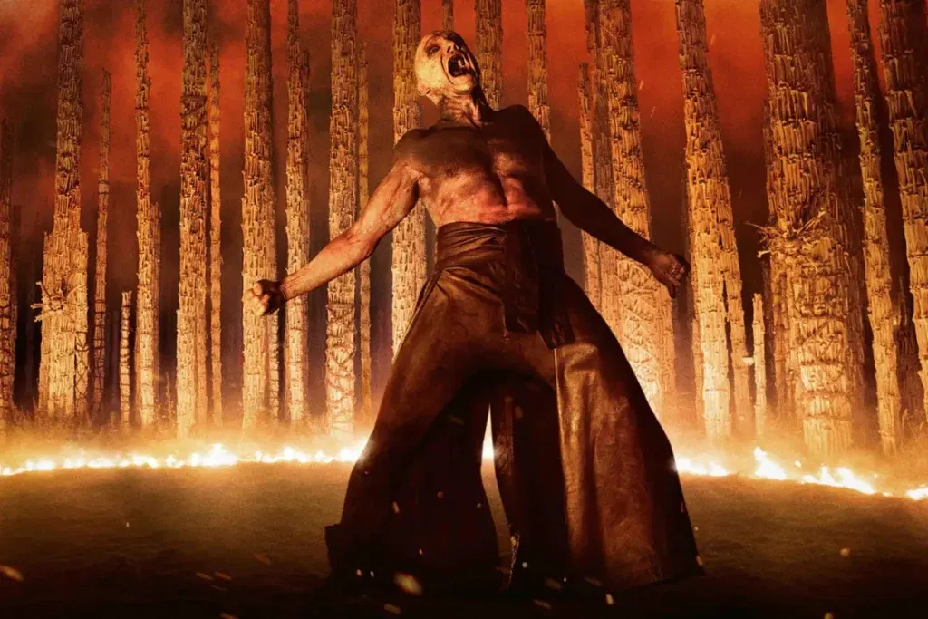 A shirtless, pale figure stands screaming amid towering bone-like structures and flames, illuminated by an intense orange glow in a nightmarish landscape.