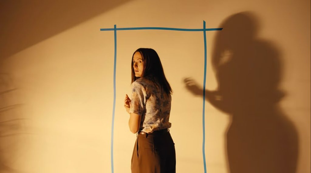 A woman with long dark hair glances back over her shoulder with a startled expression, standing before a warm beige wall. Blue tape outlines a doorframe behind her, and the shadow of a looming figure with outstretched hand is cast on the wall beside her.