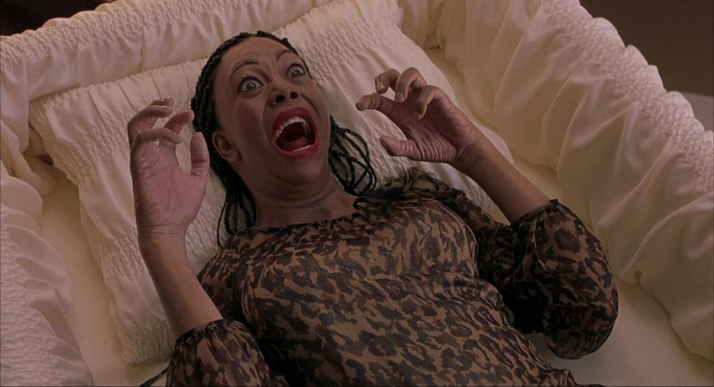 Regina Hall as Brenda Meeks lying on a bed in a leopard-print dress, screaming with hands raised in mock horror, braids splayed around her.