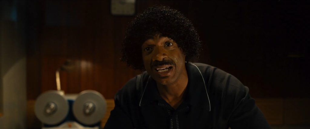Marlon Wayans as Shorty Meeks in a dark track jacket and afro hair, leaning forward mid-sentence in a dimly lit room with dumbbells in the background.