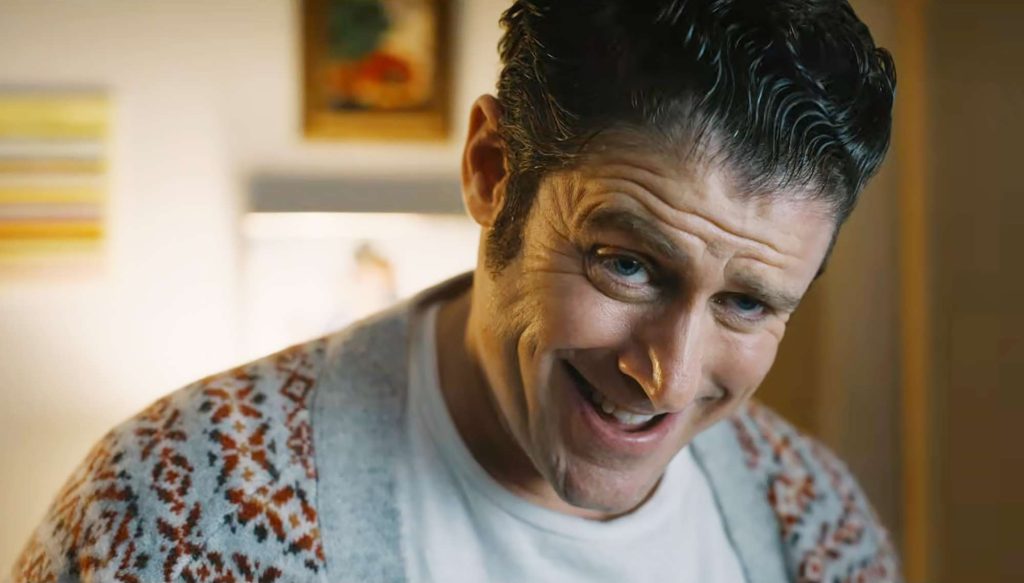 Jon Abrahams as Bobby Prinze leaning forward with a sly, unsettling grin, wearing a patterned cardigan over a white t-shirt.