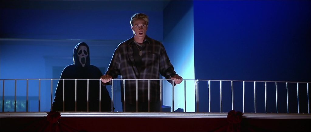 Lochlyn Munro as Greg Phillippe standing at a balcony railing with a startled expression, Ghostface lurking directly behind him in the blue-lit darkness.