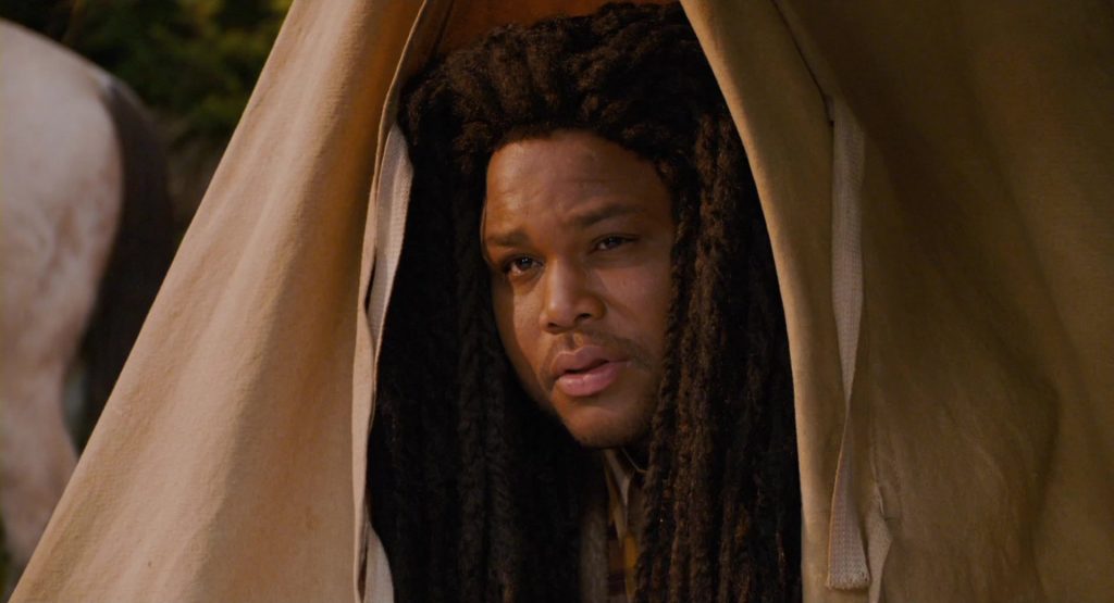 Anthony Anderson as Mahalik peering warily through a gap in a canvas tent, his face framed by long dreadlocks and a concerned expression.