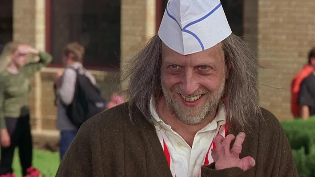 Chris Elliott as Hanson in a paper sailor hat and disheveled brown coat, grinning unsettlingly with a gnarled hand raised toward the camera.