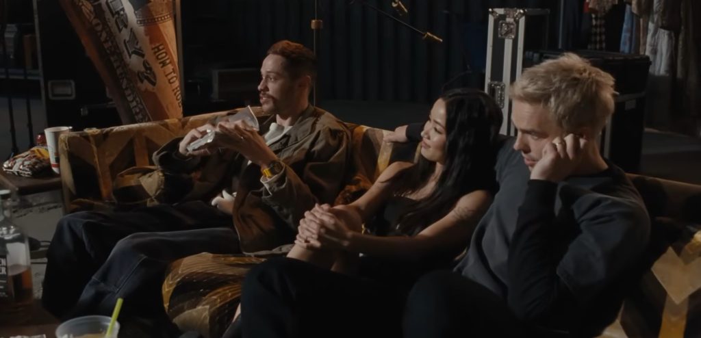 Pete Davidson, Anna Sawai, and Nicholas Hoult lounging on a worn couch in a cluttered backstage space, a "How to Rob a Bank" banner visible behind them.