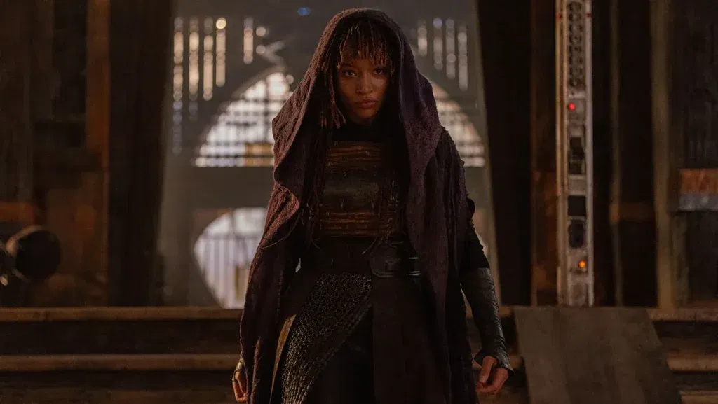 Amandla Stenberg as Mae Aniseya standing in a dimly lit corridor wearing a hooded cloak in Star Wars: The Acolyte