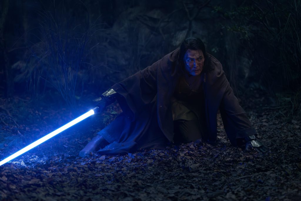 A Jedi crouches on a forest floor at night, gripping a glowing blue lightsaber as its light illuminates the surrounding leaves and casts a blue glow across the character's face in The Acolyte.