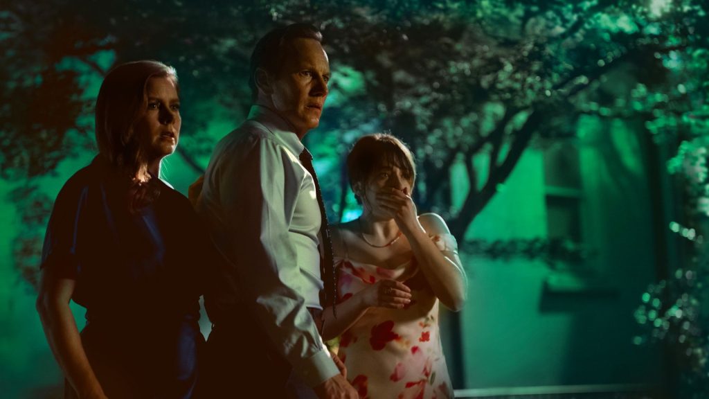 Three members of the Bowden family stand outdoors at night, staring in alarm at something off-screen as green light filters through the trees around them.