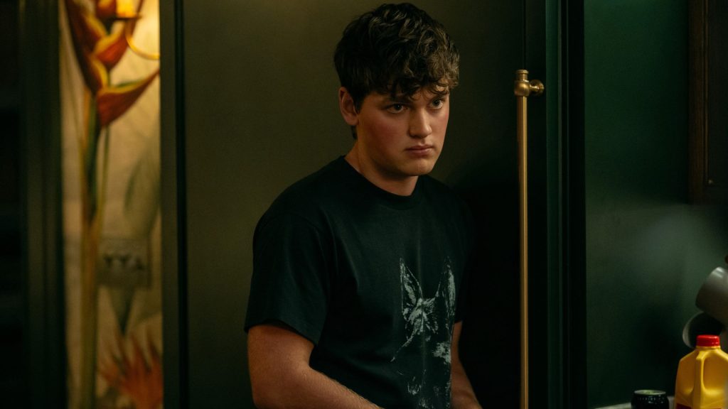 Zack wearing a black graphic T-shirt stands in a doorway, watching cautiously with a serious and guarded expression.