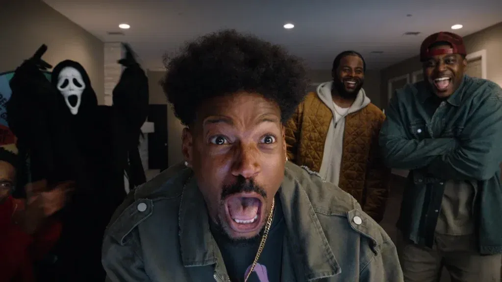 Marlon Wayans as Shorty Meeks screams in terror at the camera while a Ghostface killer looms in the background, as two laughing bystanders look on in Scary Movie 6.