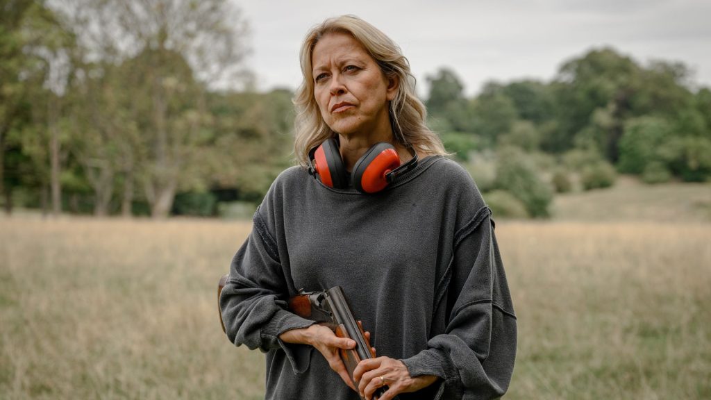 Alice and Steve Season 1 Ending Explained: Did Alice and Steve Survive the Fire? 2 Alice stands in an open field holding a shotgun, with bright orange hearing protectors around her neck and a stern, contemplative expression on her face.
