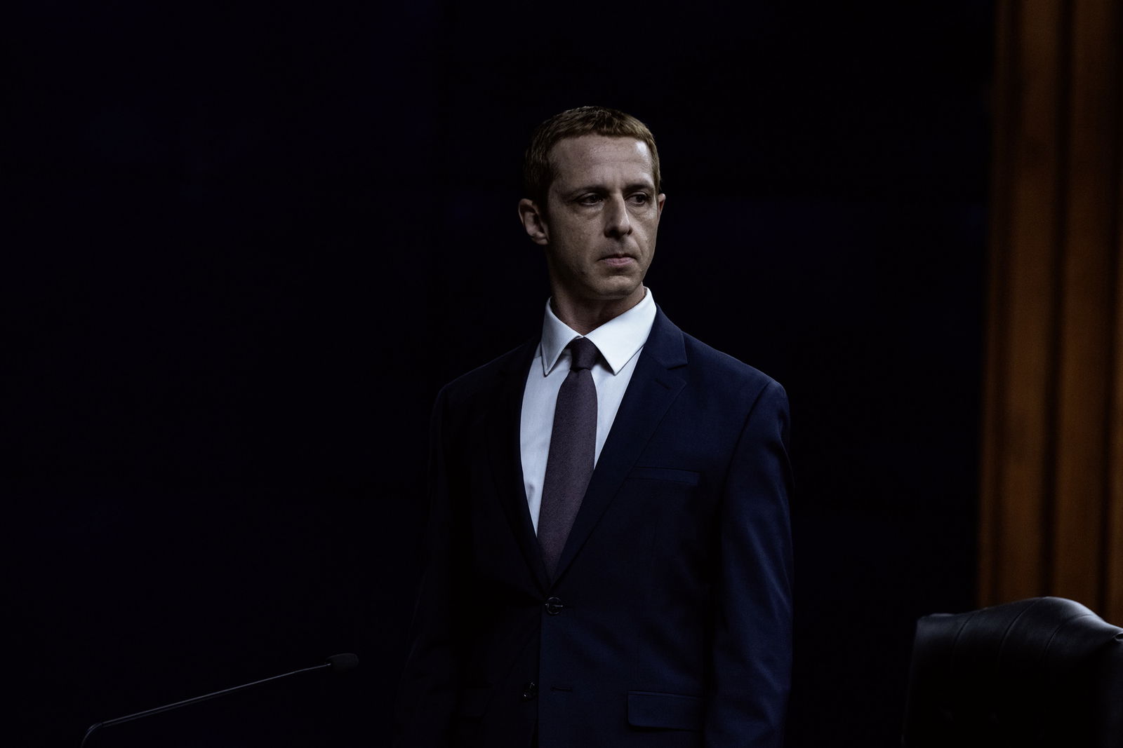 Jeremy Strong as Mark Zuckerberg in The Social Reckoning, standing in a dark room wearing a navy suit and burgundy tie