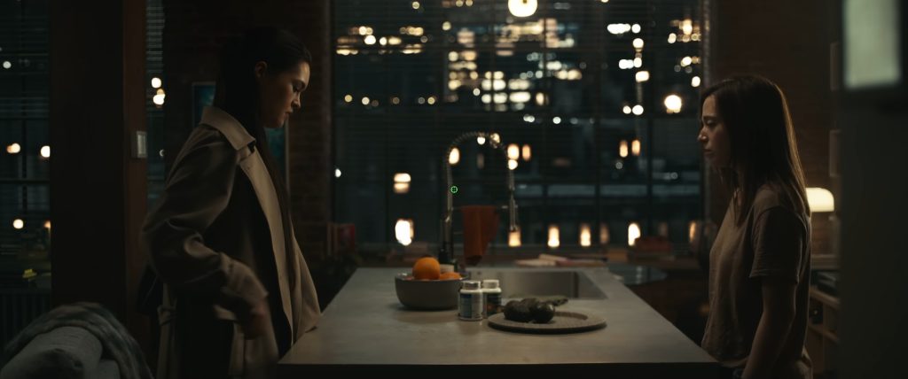 Two women face each other across a kitchen counter in a tense nighttime scene from The Social Reckoning, with a city skyline visible through the window behind them