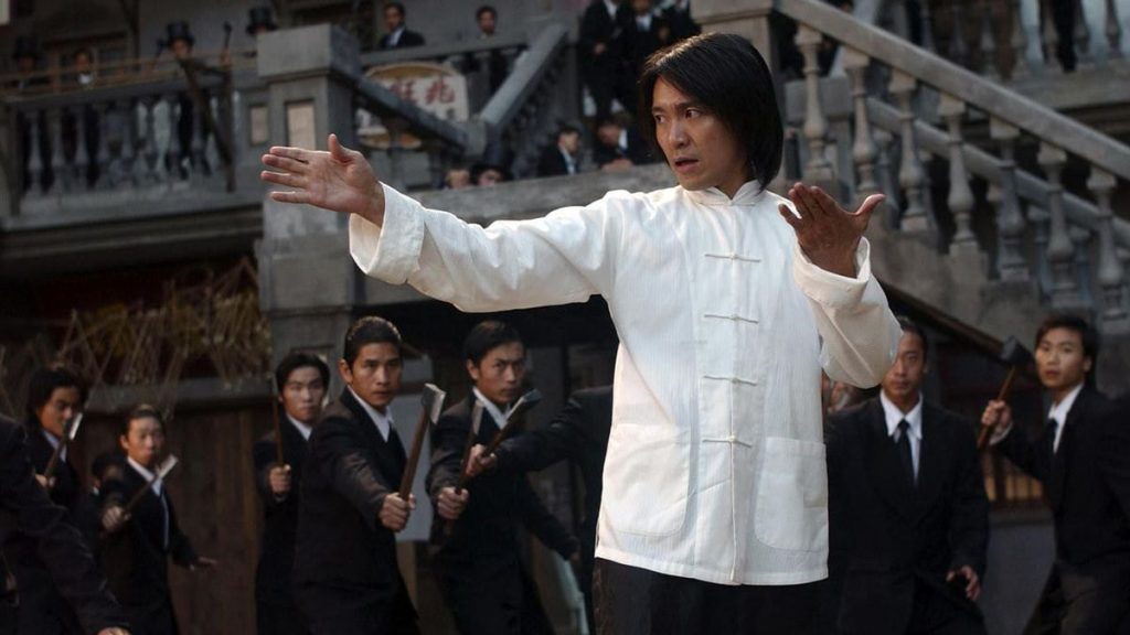 A martial artist in a white traditional Chinese tunic stands in a defensive stance at the center of a crowded courtyard while dozens of men in black suits surround him with weapons drawn in a scene from Kung Fu Hustle.